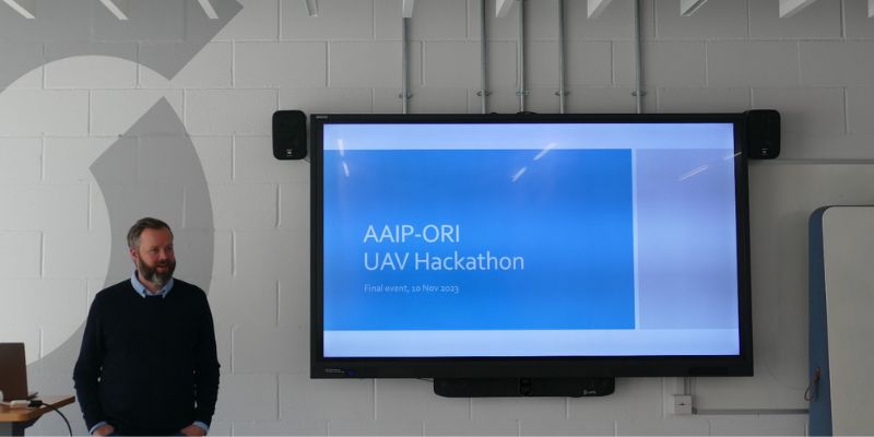 A man stands in front of a large screen. On the screen is a slide with white text on a blue background which says AAIP-ORI UAV Hackathon